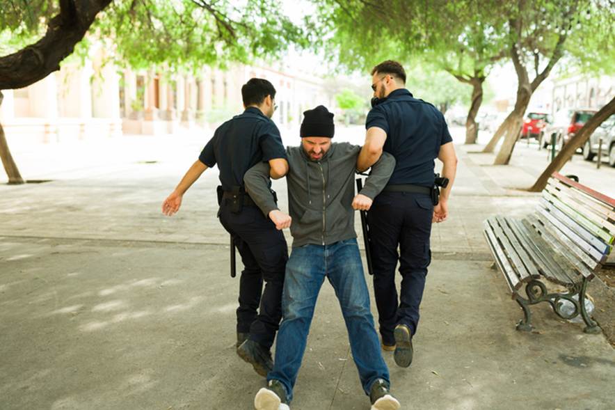 Two police officers are escorting a man wearing a beanie, gray jacket, and jeans, possibly under arrest. They are on a tree-lined street next to a park bench.